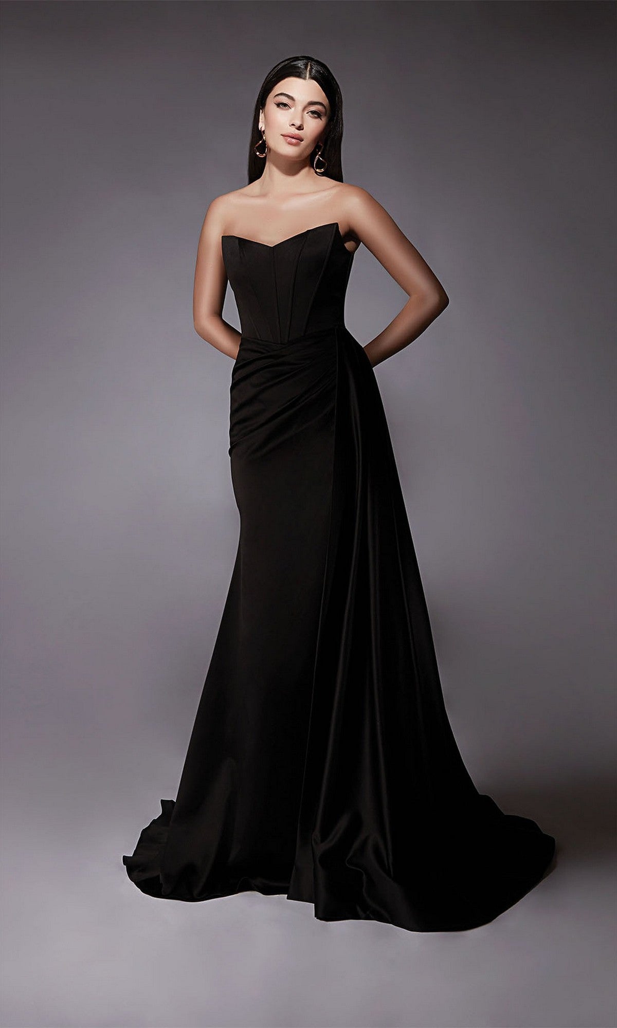 A woman poses with her hands behind her back in the Alyce 7093 long formal dress, a strapless satin gown with a fitted sheath silhouette, standing against a plain grey background.